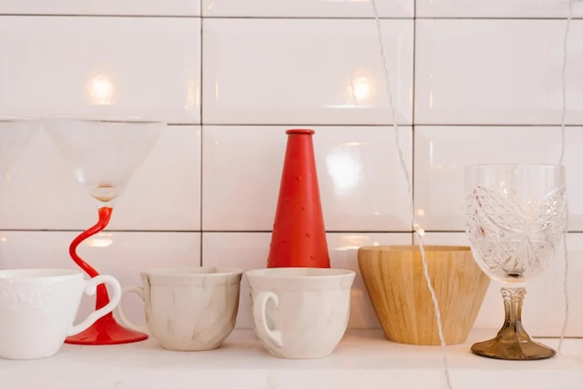 Białe porcelanowe i ceramiczne kubki, drewniane naczynia, czerwony wazon i szklanka do wina na półce kuchennej
